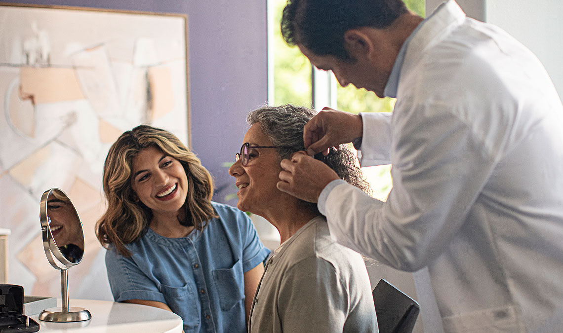 A woman watching another woman being fit with a hearing aid by a hearing professional